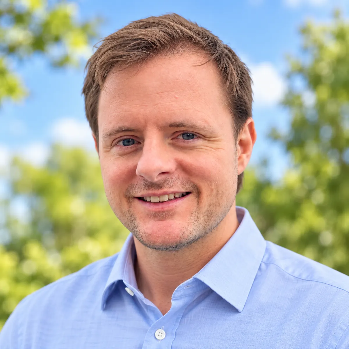 Alan Buchanan outdoors in a blue shirt, smiling, with trees behind him